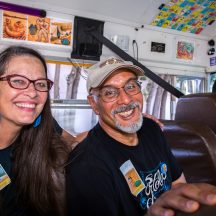Book and Wheel artists, Kate Connell and Oscar Melara are pictured on the Mexican Bus during the 5.5 Mile Road House Tour. Both are smiling. Kate has long hair and glasses.and Oscar has a ball cap on.